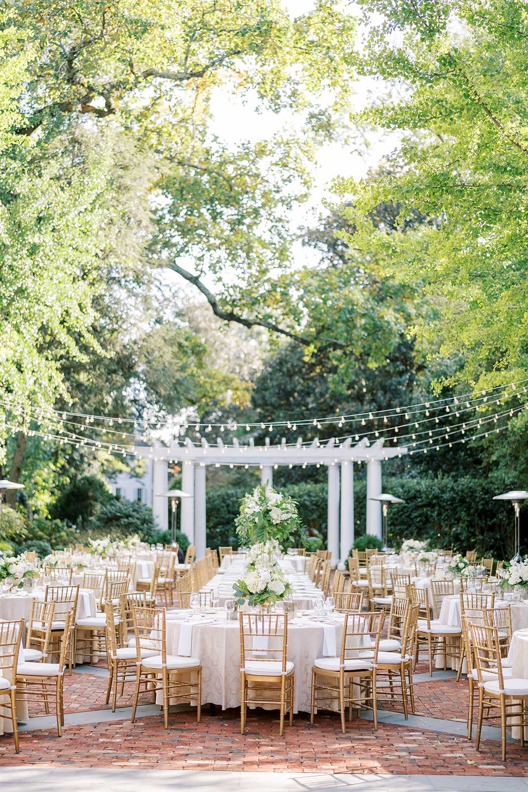 A beautifully set outdoor event space with white tables, gold chairs, and flower arrangements under string lights and a pergola.