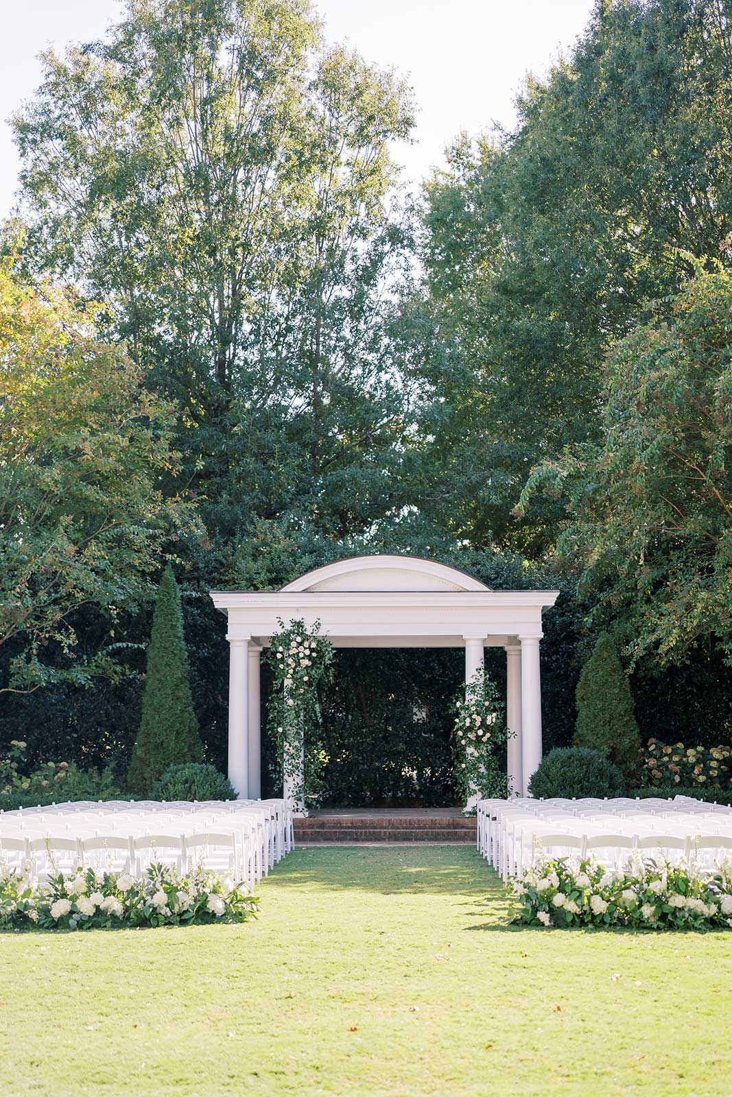 A white gazebo with flowers and white chairs set up for a wedding in a grassy area with trees.