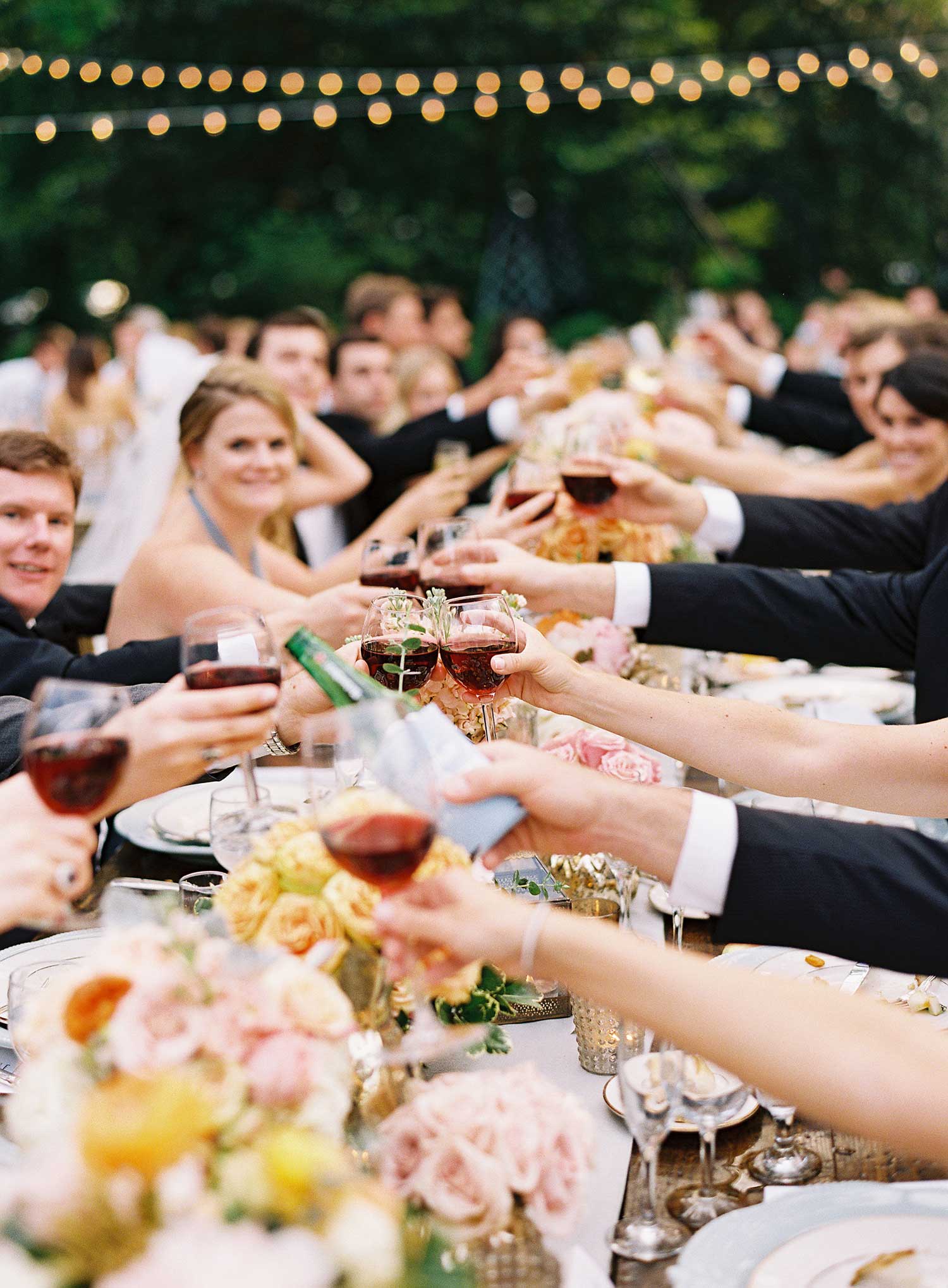 A group of people in formal attire clinking wine glasses at a long table with flowers.