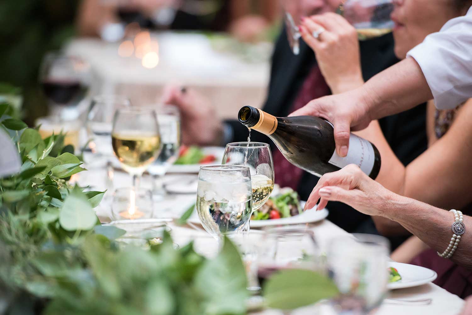 A bottle of wine is being poured into a glass on a table with salad and other glasses.